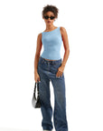 Woman in light blue one-shoulder top, wide-leg jeans, sunglasses, and white sneakers holding black bag