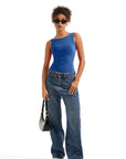 Woman in blue one-shoulder tank top, wide-leg jeans, sunglasses, and white sneakers holding black bag