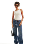 Woman in white one-shoulder top, wide-leg jeans, sunglasses, and sneakers holding black bag